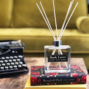 May include: A square glass diffuser with white reeds, labelled "Book Lover" with "lavender + lemon + jasmine" text. It rests on a book with a floral pattern and the title "Mansfield Park". A vintage typewriter is visible in the background.