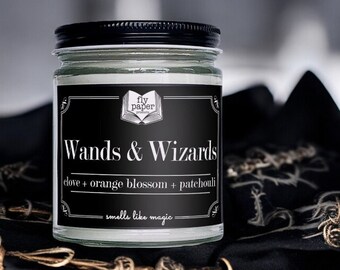 May include: A black and white candle jar with a black label that reads "Wands & Wizards" and "fly paper products". The label also includes the text "clove + orange blossom + patchouli" and "smells like magic".