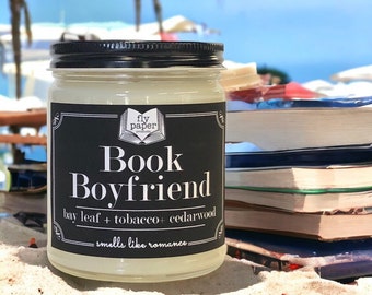 May include: A glass candle jar with a black label that reads "Book Boyfriend" and "fly paper presents". The label also says "bay leaf + tobacco + cedarwood" and "smells like romance". The candle is sitting on a beach with a stack of books behind it.