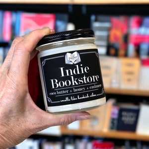 May include: A white candle jar with a black label that reads "Indie Bookstore" and "fly paper". The label also says "coco butter + honey + cassiavere" and "smells like bookish vibes".