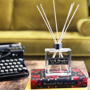 May include: A clear glass diffuser bottle with a black label that reads "Avid Reader ylang + leather + jasmine". The bottle is sitting on a stack of three books with floral patterned covers. The top book is titled "Mansfield Park JANE AUSTEN".