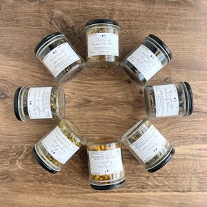 May include: A top-down view of seven glass jars with black lids, filled with dried herbs and labelled with white stickers. The labels display text such as "Anne of Green Gables" and "Through the Looking Glass". The jars are arranged in a circle on a wooden surface.