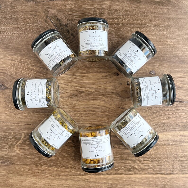 May include: A top-down view of seven glass jars with black lids, filled with dried herbs and labelled with white stickers. The labels display text such as "Anne of Green Gables" and "Through the Looking Glass". The jars are arranged in a circle on a wooden surface.