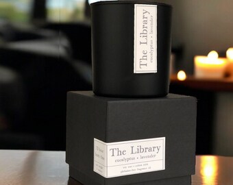 May include: Black candle in a glass jar with a black label that reads "The Library eucalyptus lavender". The candle is sitting on a black box with a white label that reads "The Library eucalyptus lavender soy wax cotton wick phthalate-free fragrance oil".