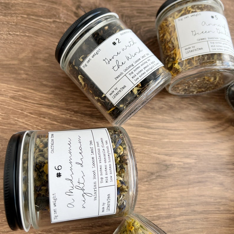 May include: A selection of glass jars filled with loose leaf tea, each with a black lid and a label detailing the tea's name and ingredients. The labels read "Gone with the Wind" and "A Midsummer Night's Dream". The jars are on a wooden surface.