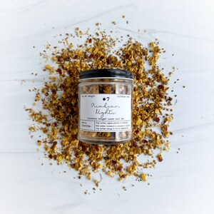 May include: A glass jar of loose leaf tea with a black lid, labelled "#7 Arabian Nights". The jar is surrounded by dried tea leaves and spices. The label indicates the tea is caffeine-free and lists ingredients like apple pieces, turmeric, and mango.