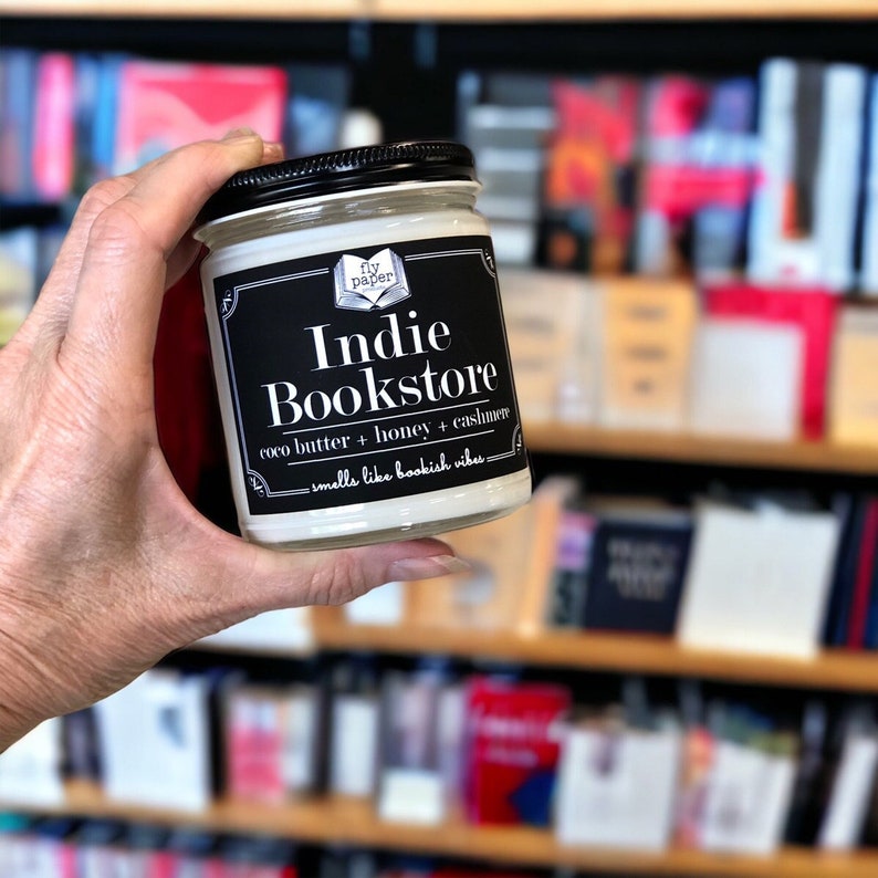 May include: A white candle jar with a black label that reads "Indie Bookstore" and "fly paper". The label also says "coco butter + honey + cassiavere" and "smells like bookish vibes".