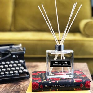 May include: A clear glass aromatherapy diffuser with white reeds, labelled "Romantasy violet + sage + saffron." It sits on a book with red floral patterns and the title "Mansfield Park." A vintage typewriter is in the background.