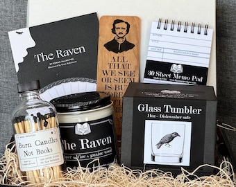 May include: A gift set featuring items with a literary theme. Includes a copy of "The Raven" by Edgar Allan Poe, a wooden bookmark, a memo pad, a glass tumbler, a candle, and a bottle of matches. The items are arranged in a black box with straw.