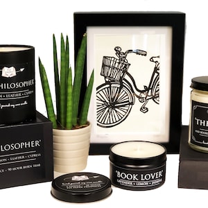 May include: Three black soy candles with labels: 'The Philosopher' with cinnamon, leather, and cypress scent, 'Book Lover' with lavender, lemon, and jasmine scent, and 'The Library' with old books, eucalyptus, and lavender scent. The candles are on a white surface with a potted plant and a framed black and white illustration of a bicycle.