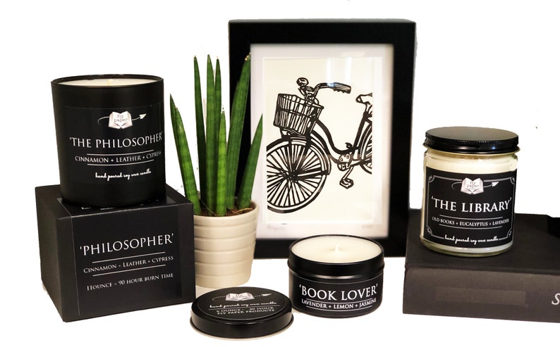 May include: Three black soy candles with labels: 'The Philosopher' with cinnamon, leather, and cypress scent, 'Book Lover' with lavender, lemon, and jasmine scent, and 'The Library' with old books, eucalyptus, and lavender scent. The candles are on a white surface with a potted plant and a framed black and white illustration of a bicycle.