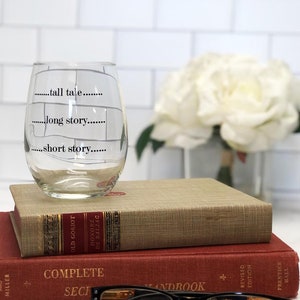May include: A clear wine glass with the phrases "tall tale," "long story," and "short story" printed on it. The glass sits atop two antique books and a pair of black-framed glasses. A bouquet of white flowers is in the background.