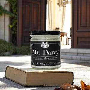 May include: A glass candle jar with a black lid and label. The label reads "Mr. Darcy" with the text "fly paper" above it. The candle is described as "cinnamon + clove + nutmeg" and "Bewitching body and soul".