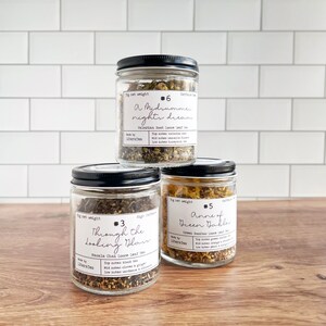 May include: Three glass jars of loose leaf tea with black lids are stacked on a wooden surface. Each jar has a label with text. The top jar reads "A Midsummer Night's Dream." The middle jar reads "Through the Looking Glass." The bottom jar reads "Anne of Green Gables."