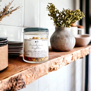 May include: A glass jar with a dark green lid containing loose leaf tea. The label reads "Arabian Nights" and "Turmeric Ginger Loose Leaf Tea." The jar sits on a wooden shelf, alongside a small vase and other decorative items.