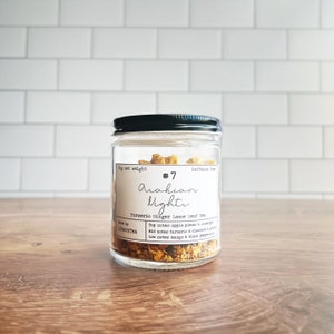 May include: A glass jar with a black lid containing loose leaf tea, labelled "Arabian Nights". The label lists ingredients including turmeric and ginger. The jar sits on a wooden surface, with a white tiled background. The tea is caffeine-free.