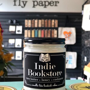 May include: A glass jar candle with a black label that reads "fly paper presents Indie Bookstore coco butter + honey + cashmere - smells like bookish vibes - "