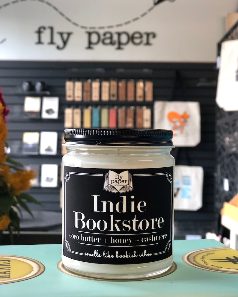 May include: A glass jar candle with a black label that reads "fly paper presents Indie Bookstore coco butter + honey + cashmere - smells like bookish vibes - "