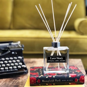 May include: A clear glass diffuser bottle with a silver cap and wooden sticks. The bottle is labelled "Love Story" with the scent notes "mahogany, amber, lavender". The diffuser is sitting on a stack of books with the titles "Mansfield Park" and "The Sun and Her Flowers".