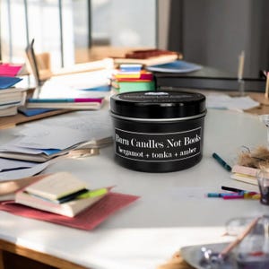 Könnte beinhalten: Eine schwarze Kerzen-Dose mit dem Text "Burn Candles Not Books" und dem Duft "bergamot + tonka + amber". Die Kerze steht auf einem Tisch mit Büchern und Papieren.