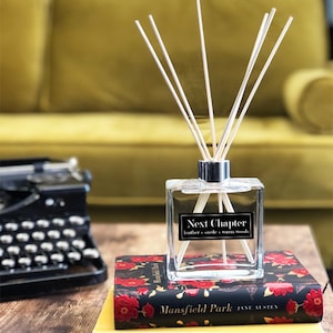 May include: A clear glass diffuser bottle with a silver cap and wooden sticks. The bottle has a label that reads "Next Chapter" with the scent description "leather suede warm woods". The diffuser is sitting on a stack of books with the titles "Mansfield Park" and "The Sun and Her Flowers".