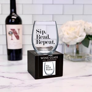 May include: A clear glass wine glass with the text "Sip. Read. Repeat." printed on it. The glass is sitting on a black box with the same text printed on it. The box is 15 ounces and dishwasher safe.