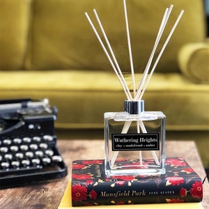 May include: A clear glass diffuser bottle with a silver cap and wooden sticks. The bottle is labelled "Wuthering Heights clay + sandalwood + amber". The diffuser is sitting on a stack of books with the titles "Mansfield Park" and "The Sun and Her Flowers".