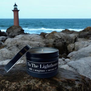 May include: A black candle tin with a white lid. The candle is labelled "To The Lighthouse" with the scent description "salt + seagrass + ocean air". The candle is sitting on a rock with a lighthouse and ocean in the background.