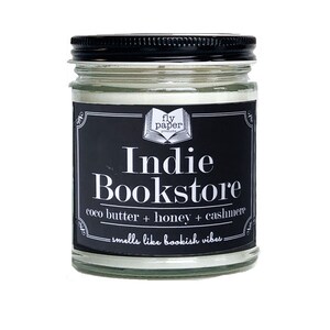 May include: A black and white candle jar with a label that reads "fly paper products Indie Bookstore coco butter + honey + cashmere -smells like bookish vibes-"