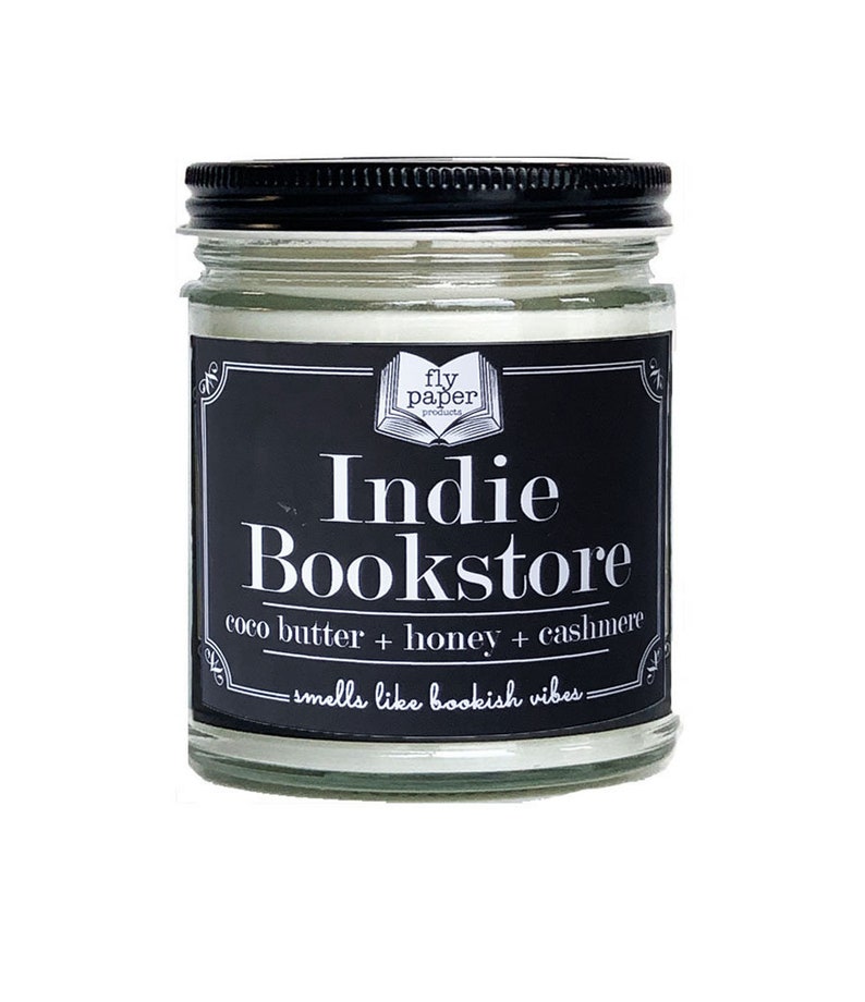 May include: A black and white candle jar with a label that reads "fly paper products Indie Bookstore coco butter + honey + cashmere -smells like bookish vibes-"