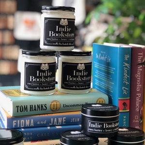 May include: A stack of books with three candles on top. The candles are labeled "Indie Bookstore" and have a scent of coco butter, honey, and cashmere. The books are titled "The Kindest Lie", "The Magnolia Palace", "Some Stories", "Uncommon Truth", "Fiona and Jane", "Call Us What We Are", and "Bertie".
