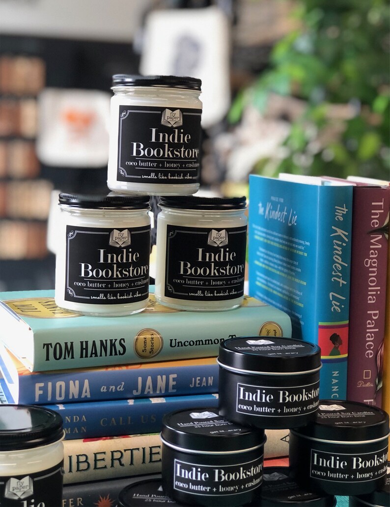 May include: A stack of books with three candles on top. The candles are labeled "Indie Bookstore" and have a scent of coco butter, honey, and cashmere. The books are titled "The Kindest Lie", "The Magnolia Palace", "Some Stories", "Uncommon Truth", "Fiona and Jane", "Call Us What We Are", and "Bertie".