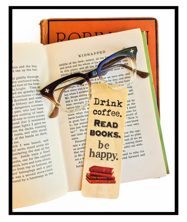 May include: A wooden bookmark with the text "Drink coffee. Read books. Be happy." and a stack of books on top. The bookmark is sitting on top of an open book with a pair of glasses on top of the book.