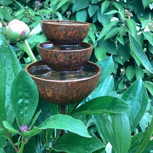 May include: A tiered, rustic-style water fountain with three bowls, each holding water. The fountain is made of a textured, brown material and is surrounded by lush green foliage and peony buds. A decorative garden accent.