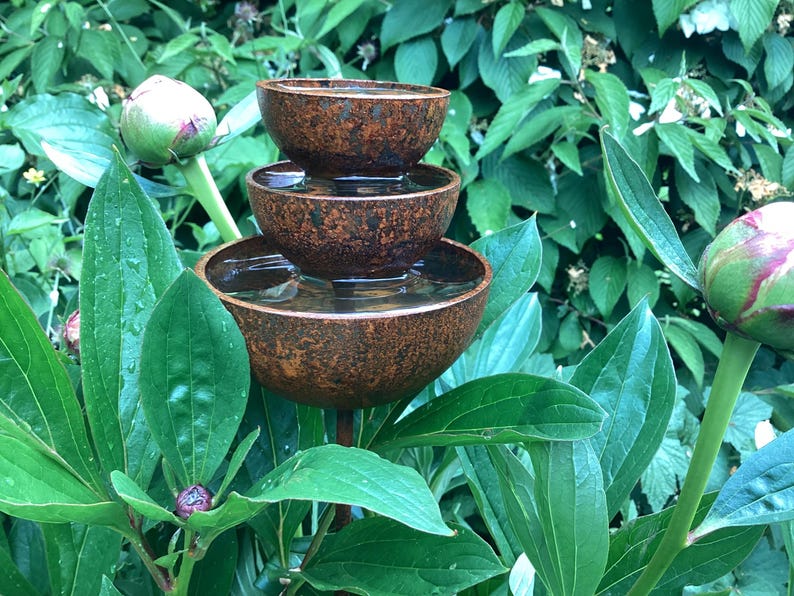 May include: A tiered, rustic-style water fountain with three bowls, each holding water. The fountain is made of a textured, brown material and is surrounded by lush green foliage and peony buds. A decorative garden accent.