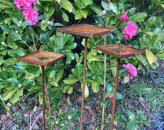 May include: Three square, rusty metal bird feeders on slender poles stand in a garden. The feeders are surrounded by green foliage and vibrant pink flowers. The feeders are a decorative garden accent.