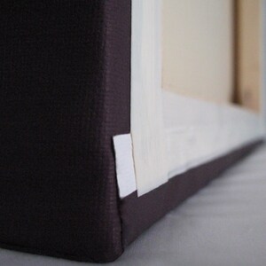 May include: Close-up of a wrapped canvas art piece. The canvas is stretched over a wooden frame and wrapped with a dark purple fabric. White tape secures the canvas to the frame.