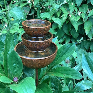 May include: A tiered, rustic-style water fountain with three bowls, each holding water. The fountain is made of a brown, textured material and is surrounded by lush green foliage. The bowls are stacked, creating a cascading effect.