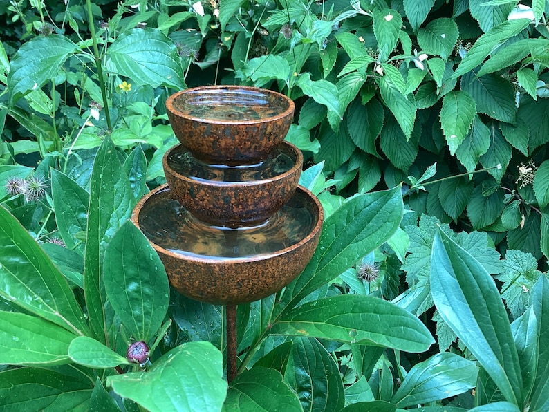 May include: A tiered, rustic-style water fountain with three bowls, each holding water. The fountain is made of a brown, textured material and is surrounded by lush green foliage. The bowls are stacked, creating a cascading effect.