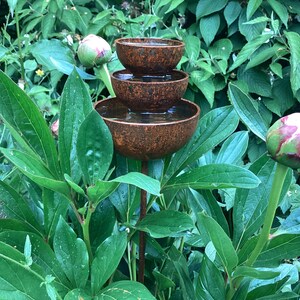 May include: A tiered, rustic metal water fountain with three bowls, filled with water, stands amidst lush green foliage. The fountain has a weathered, brown finish. A peony bud is visible in the foreground.