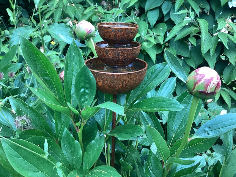 May include: A tiered, rustic metal water fountain with three bowls, filled with water, stands amidst lush green foliage. The fountain has a weathered, brown finish. A peony bud is visible in the foreground.
