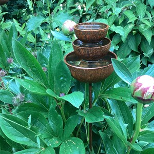 May include: A tiered, rustic-style water fountain with three bowls, set amidst lush green foliage. The bowls are a mottled brown color, and the water is visible within. The fountain is supported by a slender, dark metal rod, creating a natural garden accent.