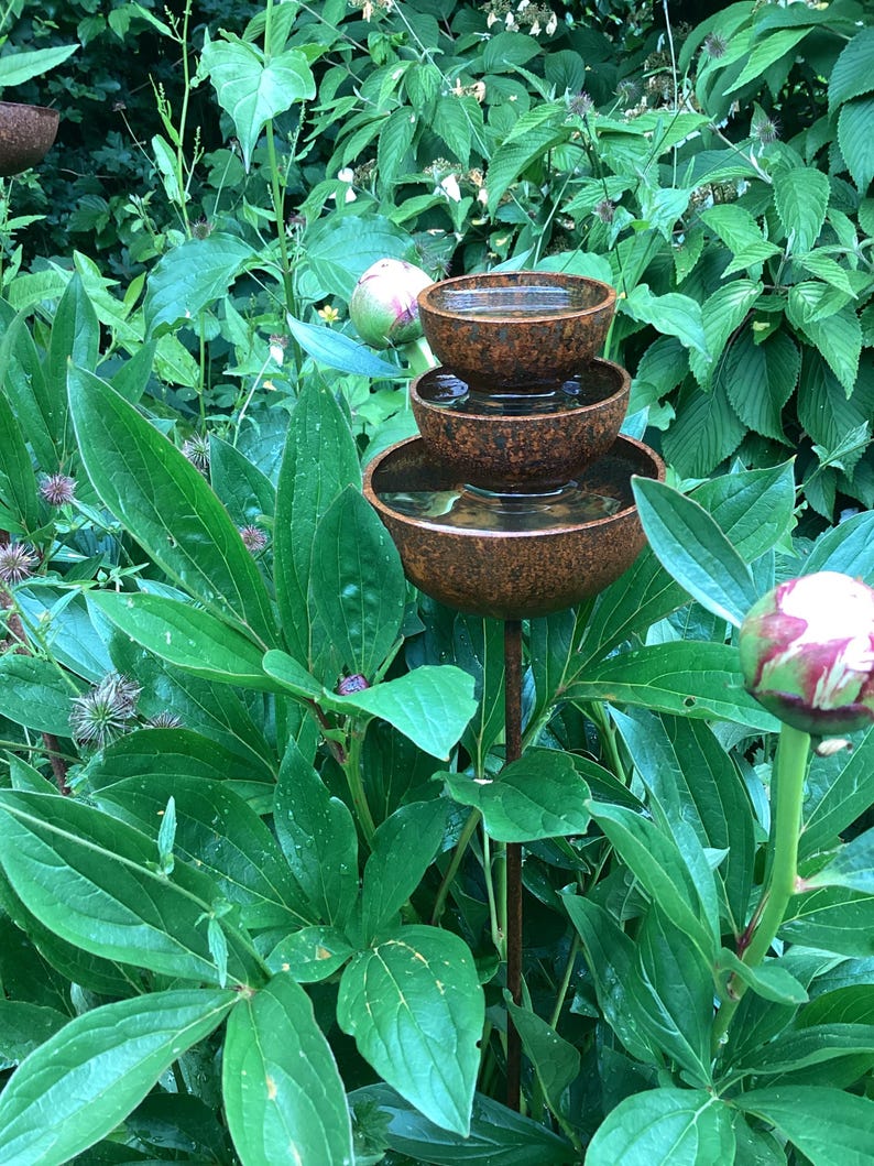 May include: A tiered, rustic-style water fountain with three bowls, set amidst lush green foliage. The bowls are a mottled brown color, and the water is visible within. The fountain is supported by a slender, dark metal rod, creating a natural garden accent.