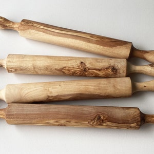 May include: Five wooden rolling pins with rounded handles are arranged on a white surface. The rolling pins are made of light and dark wood, showcasing natural grain patterns. These kitchen tools are designed for baking and pastry preparation.