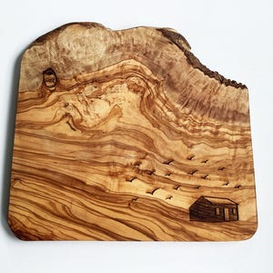May include: A live edge wooden chopping board featuring a natural grain pattern in shades of tan and brown. The surface is decorated with a wood-burned illustration of a small cabin and a flock of birds flying across the sky above the structure.