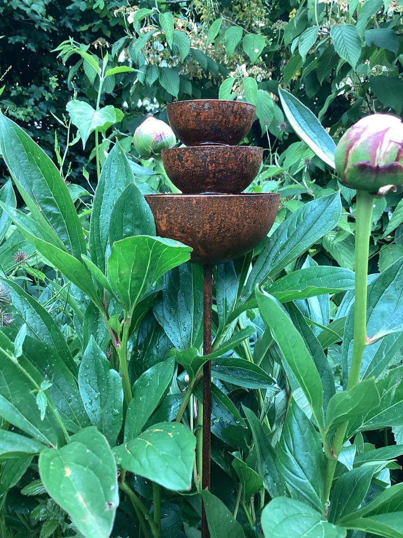 May include: A tiered, rustic metal garden fountain with three bowls of decreasing size. The fountain is set amidst lush green foliage, including large leaves and unopened flower buds. The metal has a weathered, brown patina.
