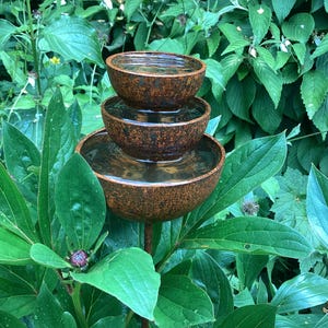 May include: A tiered outdoor water fountain with three brown, bowl-shaped basins. The fountain is filled with water and set amidst lush green foliage. The fountain is made of a textured, weathered material.