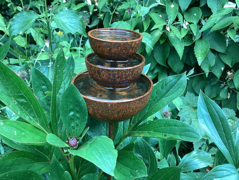 May include: A tiered outdoor water fountain with three brown, bowl-shaped basins. The fountain is filled with water and set amidst lush green foliage. The fountain is made of a textured, weathered material.