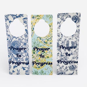 Może przedstawiać: Trzy wieszaki na drzwi z kwiatowymi wzorami i napisem "Prayer In Progress". Każdy wieszak ma otwór u góry. Wzory obejmują niebiesko-białe wzory kwiatowe oraz żółto-niebieski wzór.