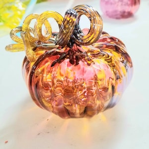 May include: A hand-blown glass pumpkin sculpture featuring a ribbed body with a gradient of pink, orange, and yellow hues. The top of the pumpkin is adorned with a clear, spiraled glass stem and two curled, amber-colored glass tendrils.